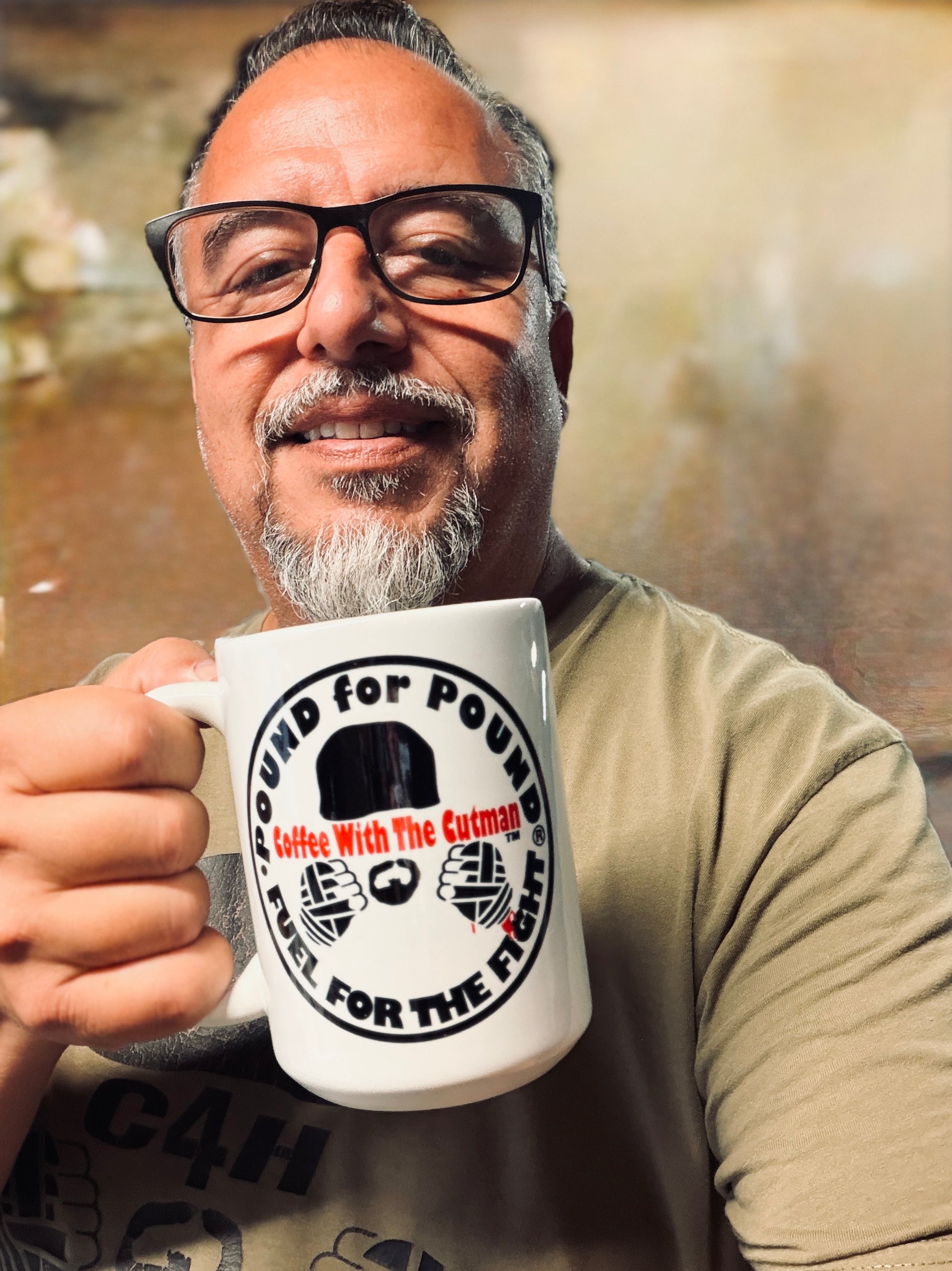 A smiling man with glasses and a gray beard holds up a white ceramic mug with a black and red graphic that reads 'Pound for Pound Coffee With The Cutman' and 'Fuel FOR THE FIGHT'.